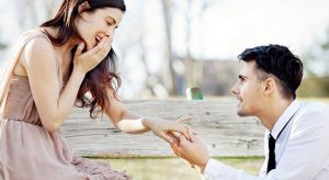 A man is holding a woman's hand while sitting on a bench, showcasing a perfect diamond engagement ring.