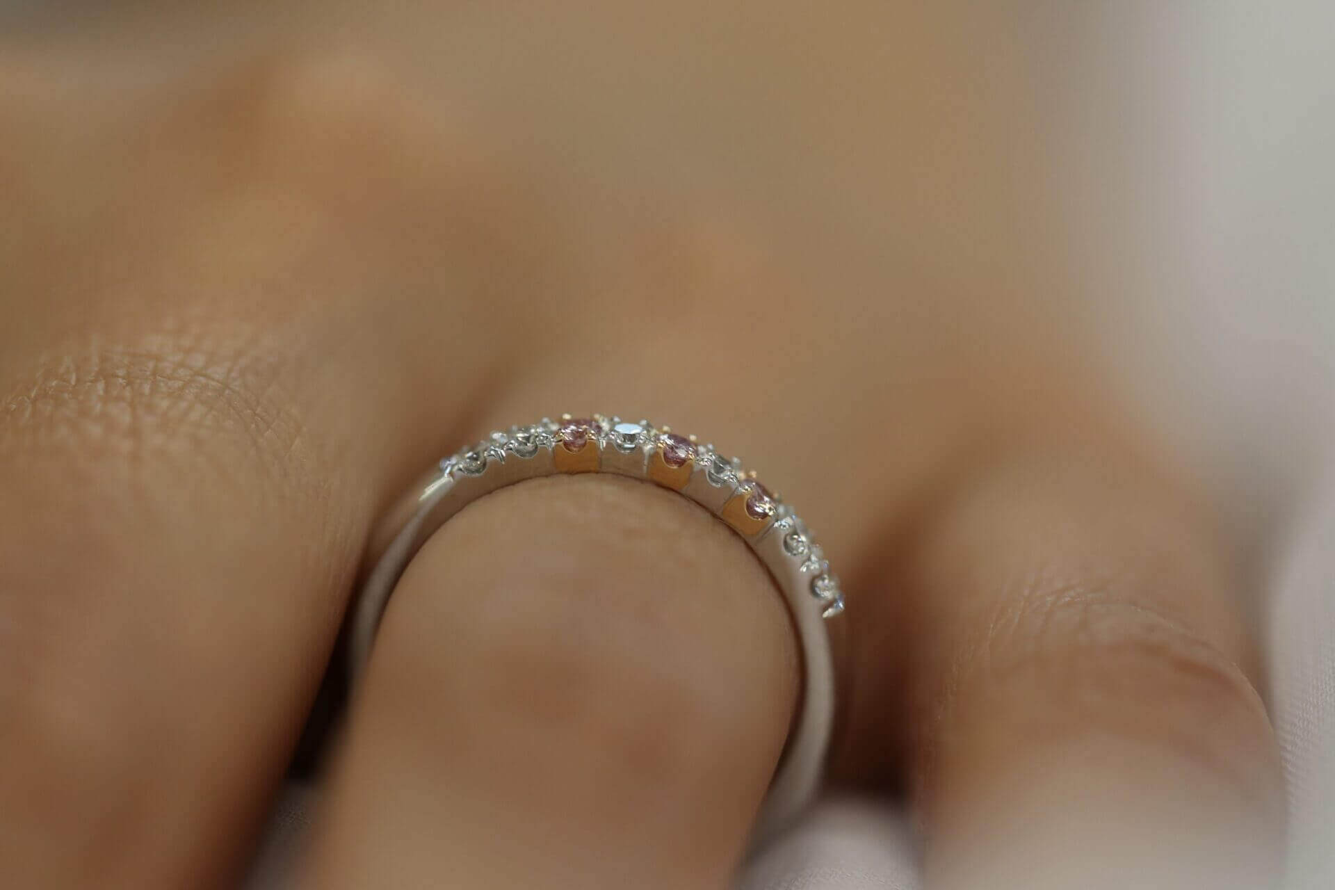 A woman's hand adorned with a White and Pink Diamond Wedding Ring C573 glimmers in the light, showcasing its exquisite beauty.
