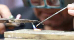 A person wearing safety glasses is carefully using soldering tools to work on a small ring on a table, ensuring there are no ring resizing mistakes.