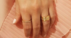 A close-up of a person's hand with three gold rings, each adorned with diamonds, on their fingers. The nails are painted white, and the background is a pink, pleated fabric. This elegant display could serve as an inspiring example in any guide to buying engagement rings.