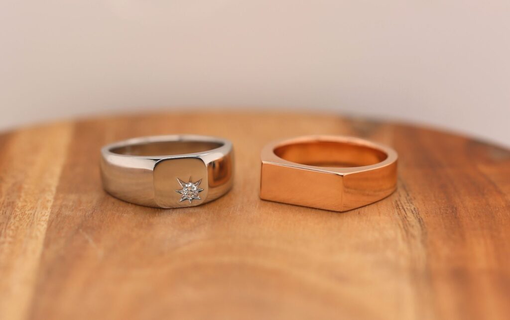 Two men's signet rings on a wooden surface: one is a white gold men’s wedding band with a star-shaped design and a small diamond, the other is a plain rose gold band.