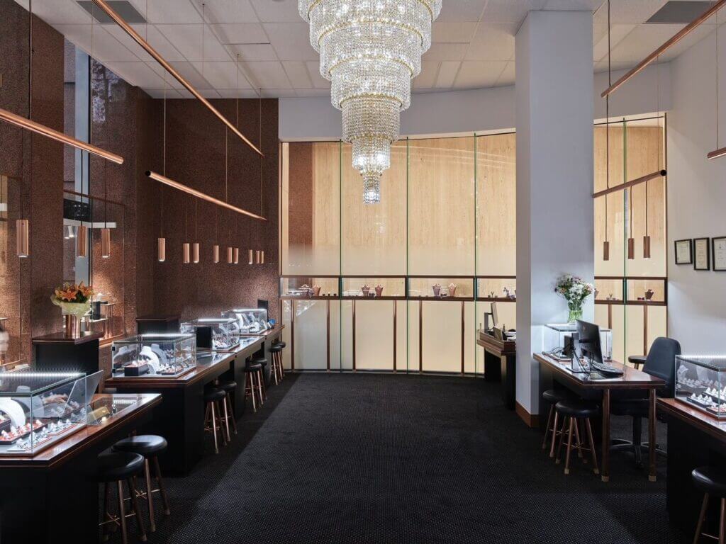 A well-lit Melbourne boutique interior with display cases showcasing handmade jewelry, brown wall panels, a large chandelier, and a black carpeted floor. There are stools at each display case and a flower vase on a desk.