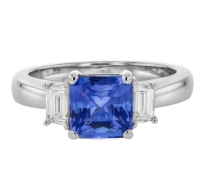 A white gold ring features a large, square cushion cut blue sapphire gemstone in the centre, flanked by two smaller, baguette white gemstones, in a Trilogy Diamond Engagement Ring style.