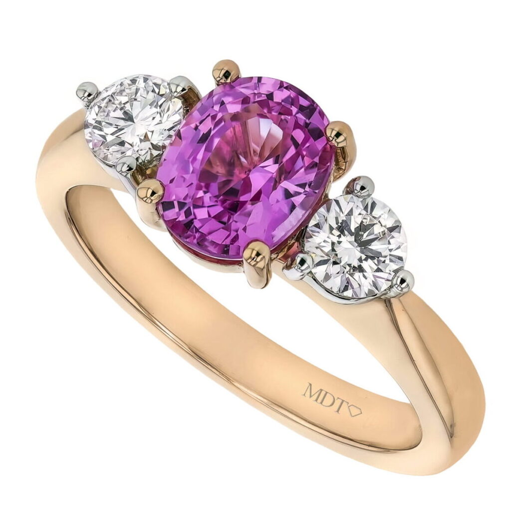A rose gold ring with a central oval pink sapphire gemstone flanked by two round white diamonds, epitomizing the elegance found in Trilogy Diamond Engagement Rings.