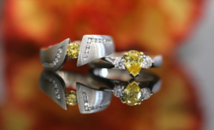 A close-up of two yellow diamond rings with small surrounding diamonds displayed on a reflective surface, set against a blurred orange and yellow background.