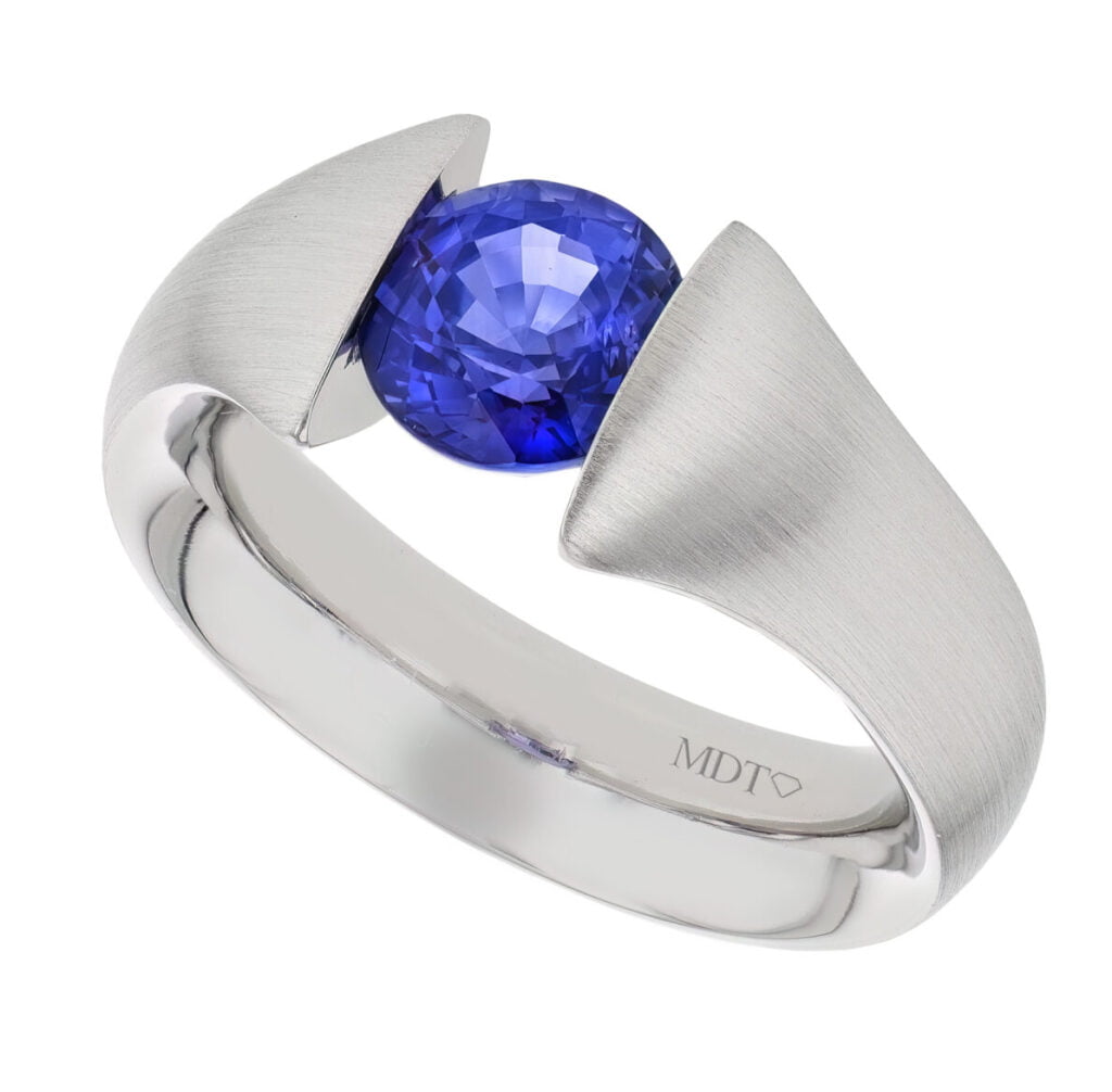A white gold tension set ring with a blue Ceylon sapphire in the centre and the inscription "MDT" on the band.