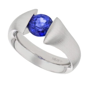A white gold tension set ring with a blue Ceylon sapphire in the centre and the inscription "MDT" on the band.
