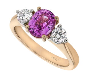A gold ring featuring an oval-cut pink sapphire flanked by two smaller round diamonds. The band, engraved with "MDT" is a stunning addition to any collection of sapphire engagement rings in Melbourne.