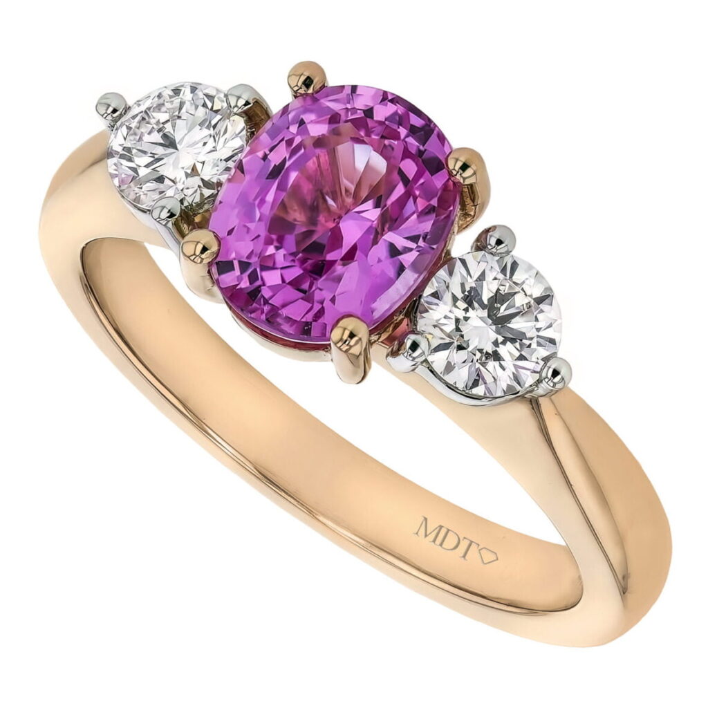 A gold ring featuring an oval pink sapphire flanked by two smaller round diamonds. The band, engraved with "MDT" is a stunning addition to any collection of sapphire engagement rings in Melbourne.