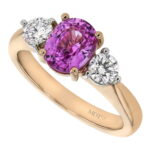 A gold ring featuring an oval pink sapphire flanked by two smaller round diamonds. The band, engraved with "MDT" is a stunning addition to any collection of sapphire engagement rings in Melbourne.