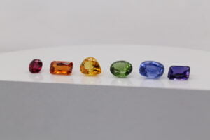 A row of six differently colored gemstones, including red, orange, yellow, green, blue, and purple, displayed on a white surface against a neutral background. These vibrant stones are perfect for creating stunning coloured sapphire engagement rings in Melbourne.