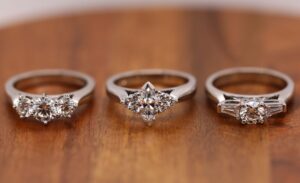 Three white gold diamond trilogy rings are displayed on a wooden surface. Each ring features a unique diamond arrangement, epitomizing the allure of the best diamond rings. The image includes the text "MDT DESIGN Individually designed, hand-made jewellery.