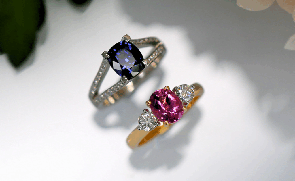 A photo of two sapphire engagement rings, one split band blue sapphire and diamond engagement ring and one trilogy engagement ring with a pink sapphire in the centre and two white diamonds either side.