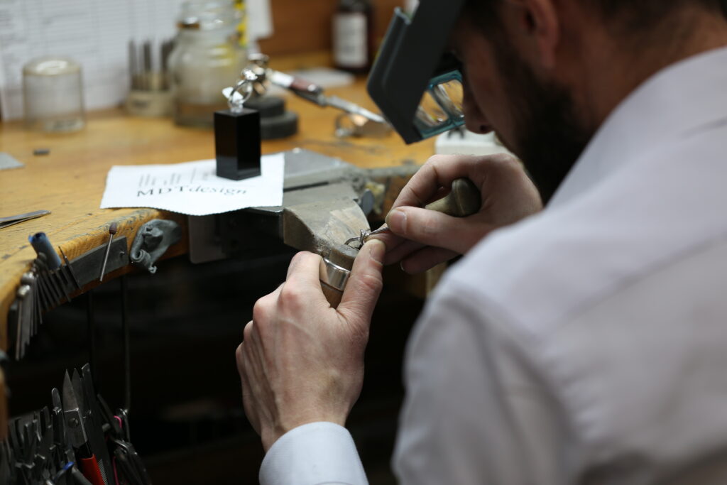 Close-up of a skilled jeweller working meticulously on a fine jewellery piece inside a professional workshop, using precision tools under focused lighting.