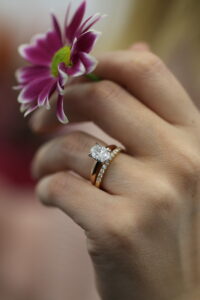 Close-up of a hand holding a pink flower wearing an MDT DESIGN rose gold oval diamond engagement ring paired with a diamond wedding band, showcasing handmade fine jewellery craftsmanship in Melbourne.