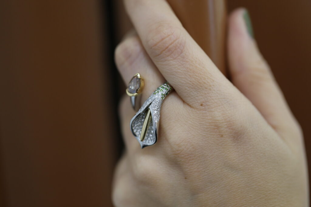 Close-up of a hand wearing MDT DESIGN’s award-winning Arum ring, a lily-inspired design featuring white and yellow gold, pavé diamonds, and green gemstone detail.