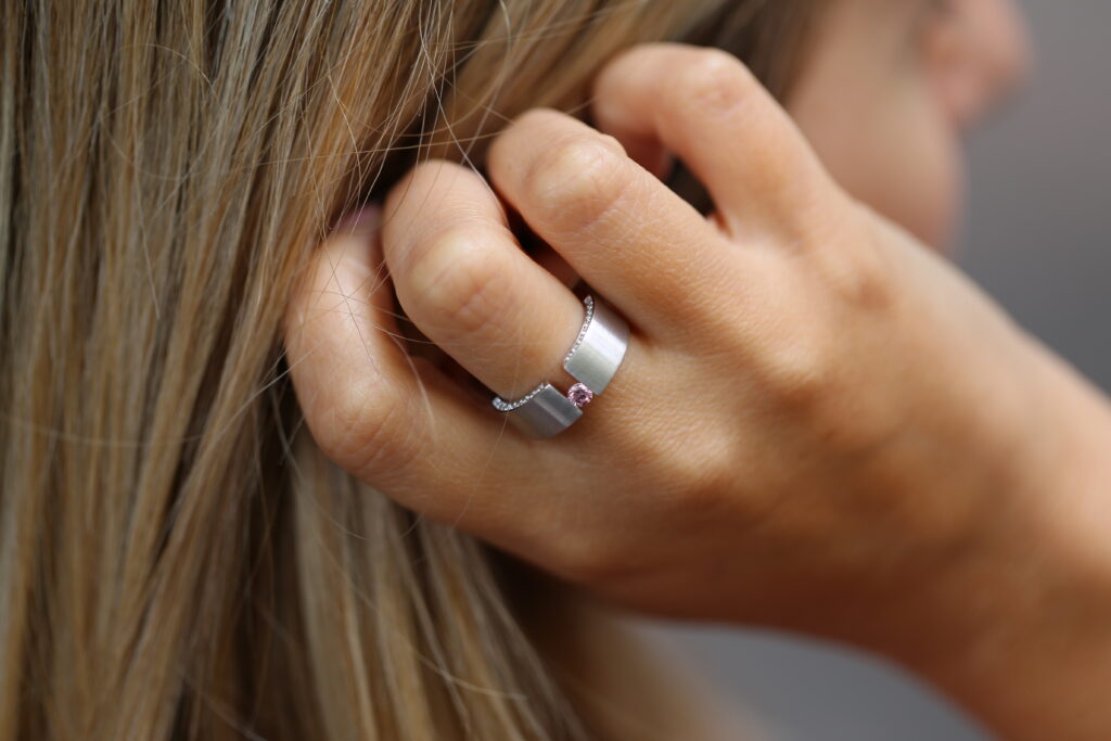 Hand pushing back hair- showcasing a unique design handmade tension set ring featuring a pink diamond and diamonds set into the side of the band.