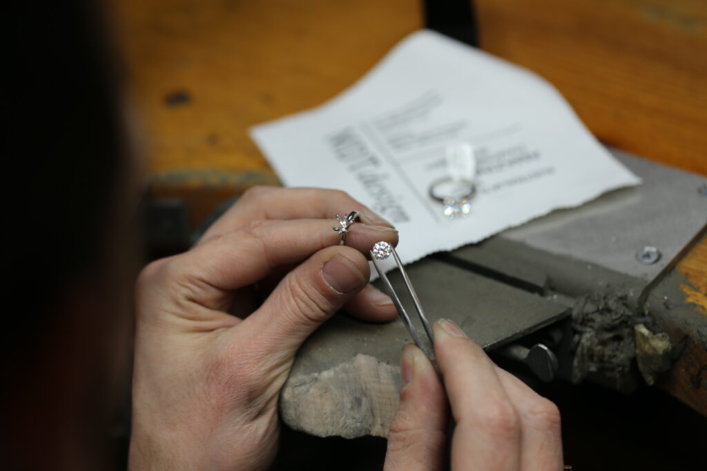 Over the shoulder of a jeweller- we see a round brilliant cut certified diamond held in tweezers next to a handmade engagement ring setting.