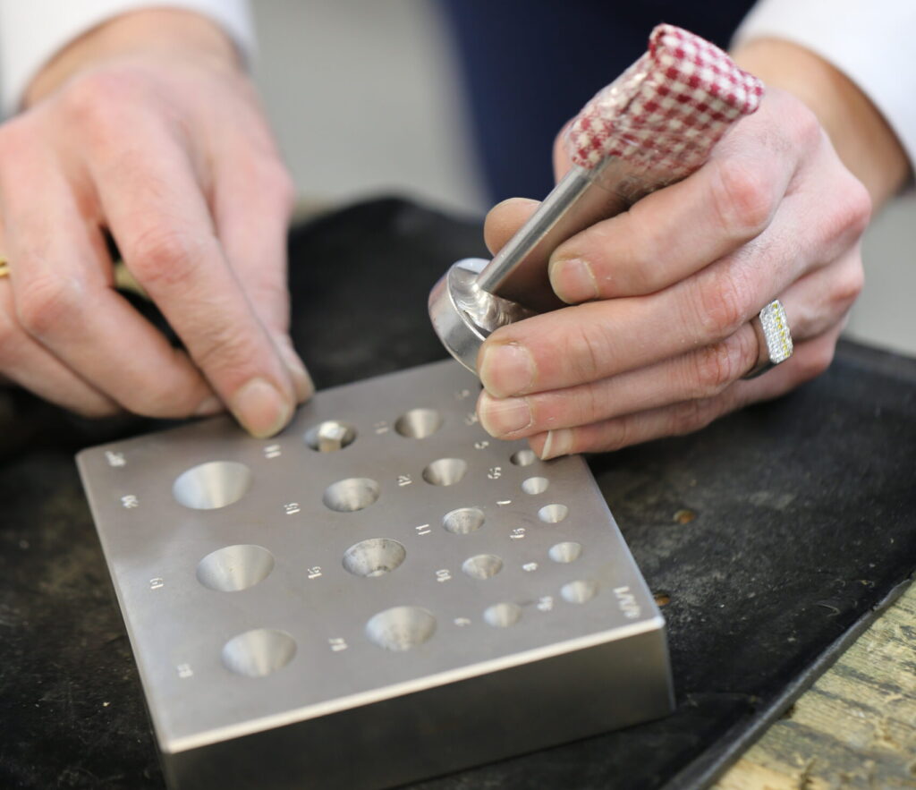 Jeweller working in a workshop- photo of hands holding a small piece of metal, hammering it into setting moulds.