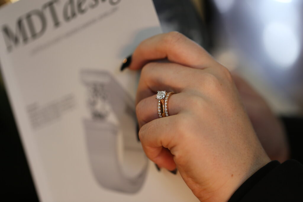 Hand wearing a rose gold claw-set diamond engagement ring with a matching claw set diamond wedding band, photographed in front of an MDT DESIGN magazine.