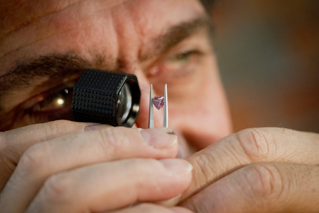MDT DESIGN DIRECTOR Nicholas Theochari holds a eye loupe to his eye magnifying a small heart cut pink diamond