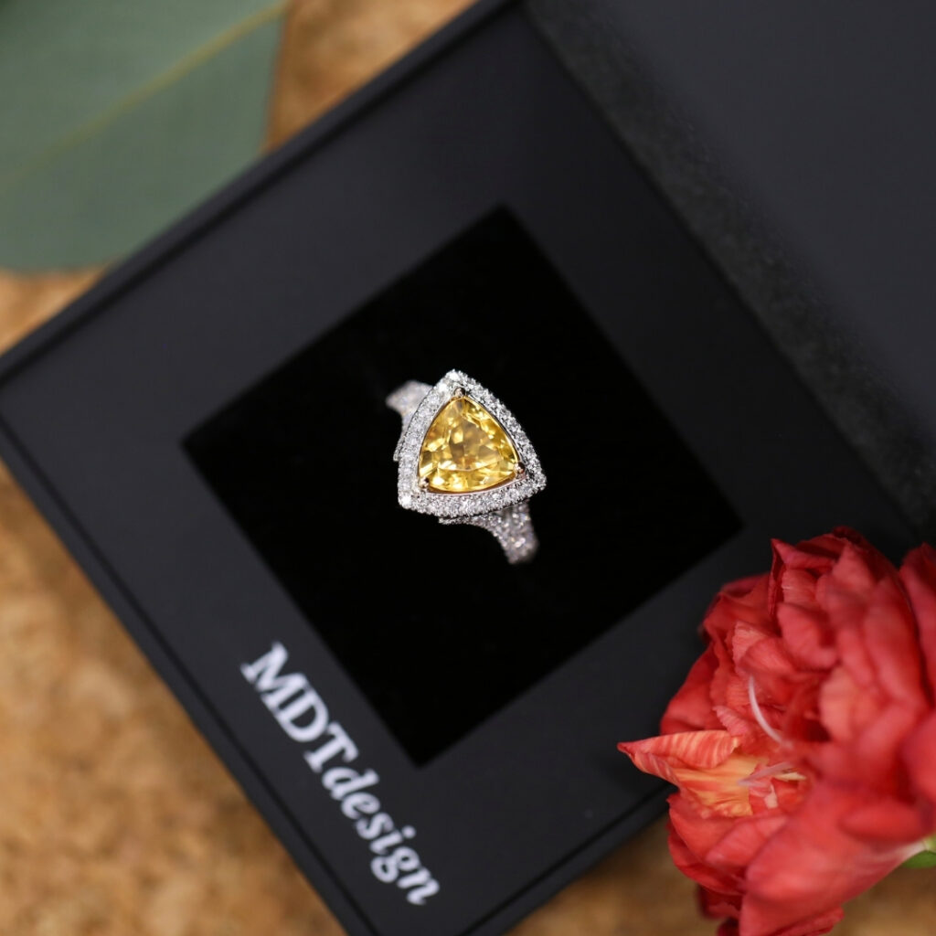 Close-up of a handmade white gold ring featuring a triangular-cut citrine centre stone, surrounded by a halo of small diamonds, displayed in an MDT DESIGN ring box beside a red flower.