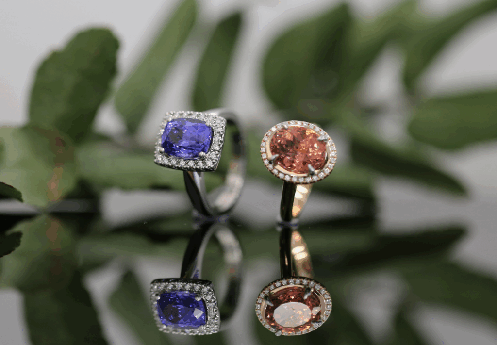 Two handmade halo rings displayed on a reflective surface - one featuring a blue tanzanite surrounded by white diamonds in white gold, and the other an oval pink tourmaline encircled by diamonds in rose gold, with soft green foliage in the background and MDT DESIGN branding.