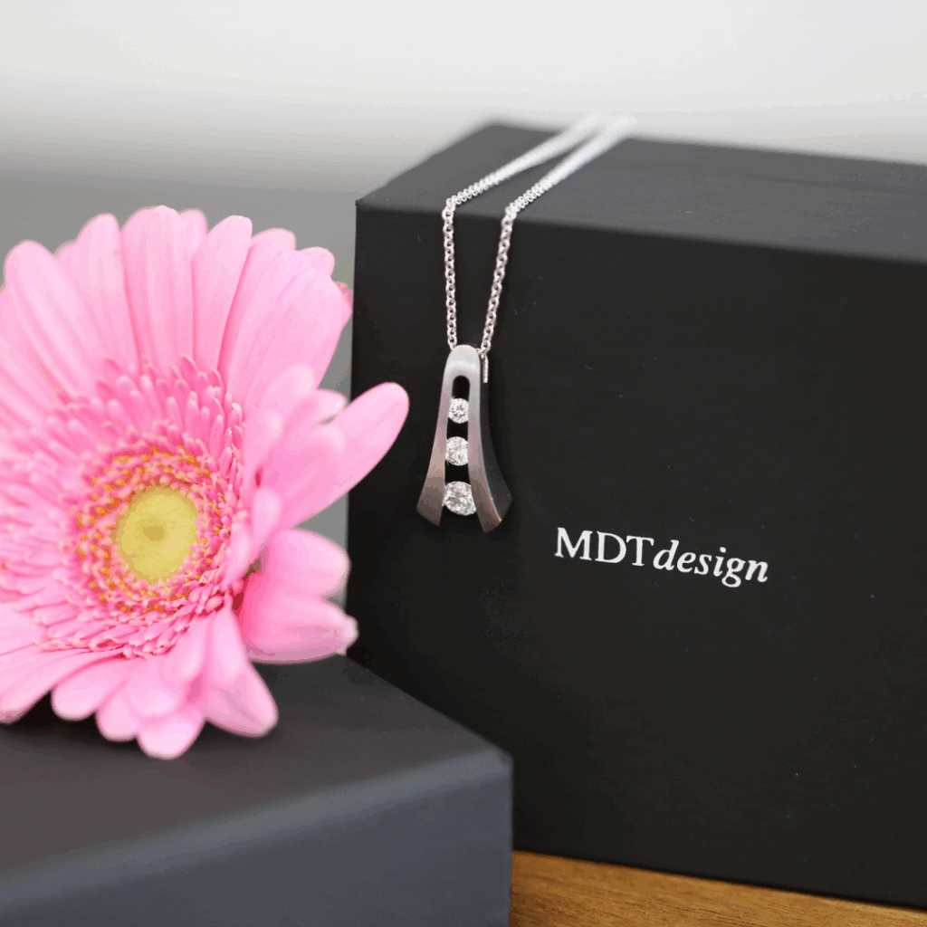 Handmade white gold split-band pendant featuring three natural white diamonds tension set vertically, suspended on a fine chain and displayed against an MDT DESIGN box beside a pink flower.
