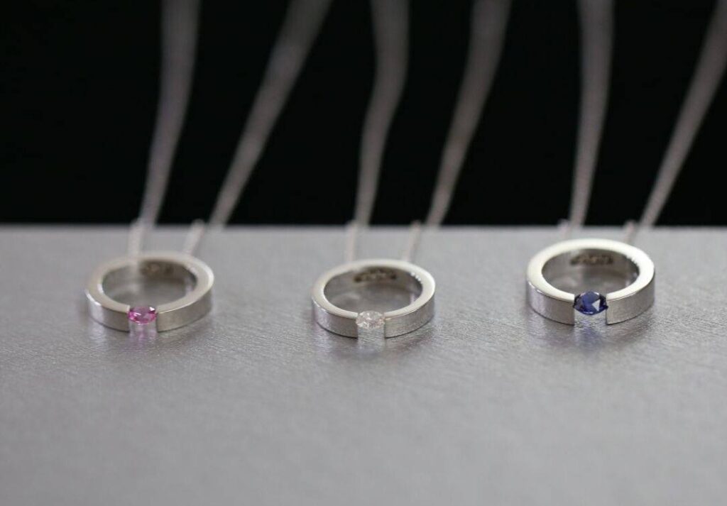 Three handcrafted white gold tension-set pendants on fine chains, each featuring a single gemstone — pink sapphire, white diamond, and blue sapphire — displayed on a brushed metal surface with MDT DESIGN branding.