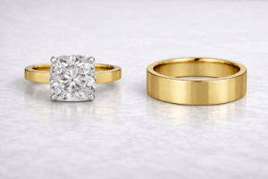 Render inspired by Ada Nicodemou’s engagement ring, featuring a square cushion-cut diamond in white claws on a flat yellow gold band, paired with a thick flat-profile yellow gold men’s wedding ring.