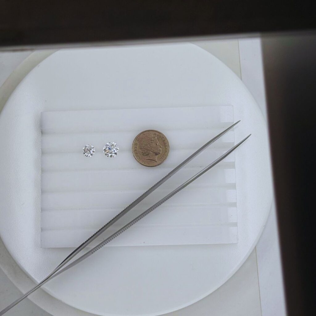 Size comparison of a 1 carat and 2 carat round brilliant cut diamond placed beside an Australian 5 cent coin, shown on a white gem tray with jeweller’s tweezers for scale