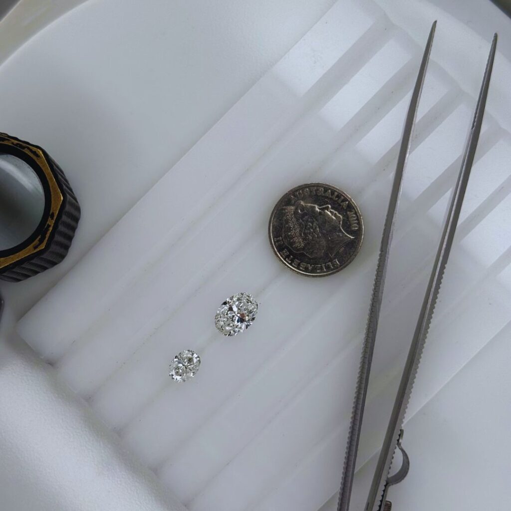 Top-down view of a 1 carat and 2 carat round brilliant cut diamond displayed on a white jeweller’s tray, positioned beside an Australian 5 cent coin and precision tweezers for size comparison.