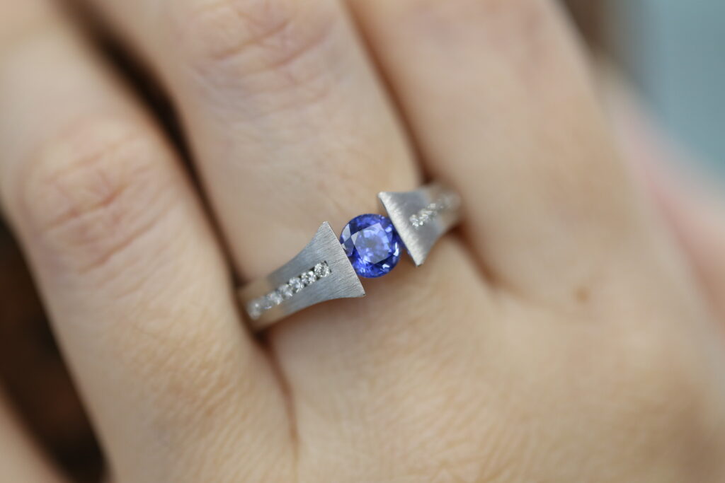 Close-up of a handmade tension set engagement ring featuring a vivid round blue sapphire suspended between brushed white metal settings, with small accent diamonds set into the band, worn on the hand.