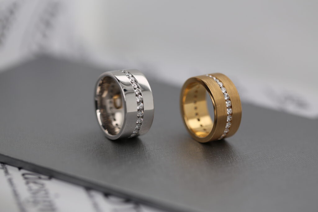 Two wide men’s diamond rings in white and yellow gold, featuring rows of round brilliant diamonds and contrasting polished and brushed finishes.