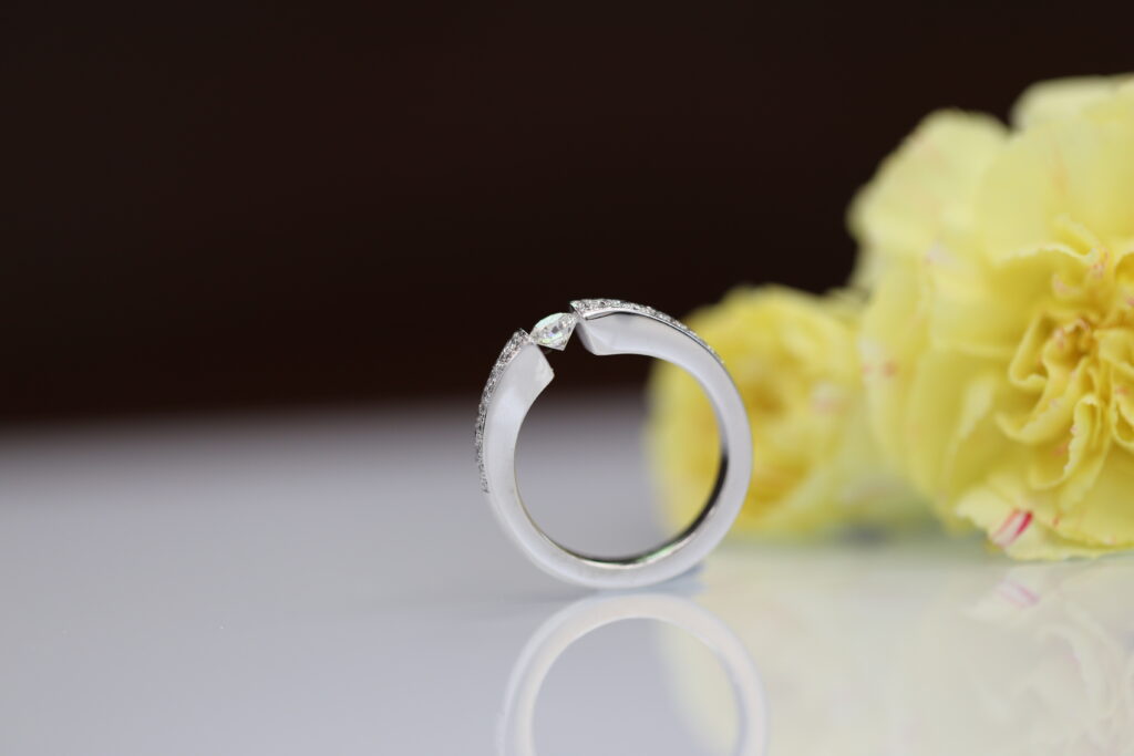 White gold tension set diamond ring with a diamond-set band, photographed beside yellow flowers.