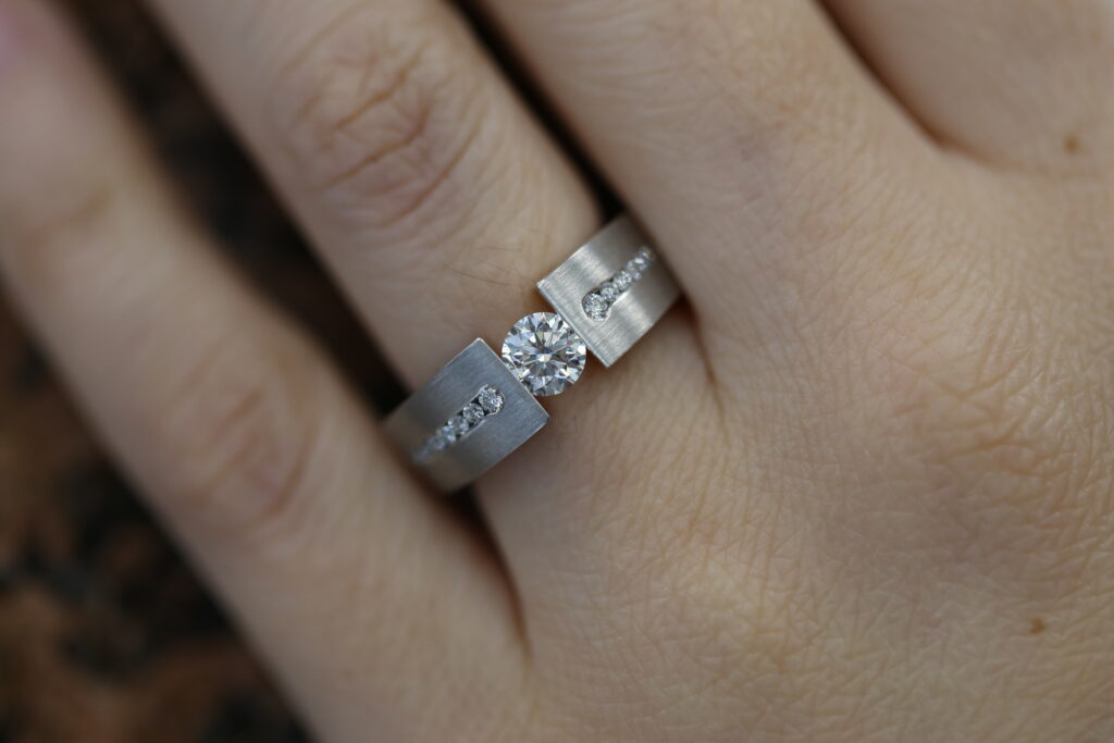 Tension set diamond ring worn on the hand, with a round brilliant centre diamond and diamond-set shoulders.